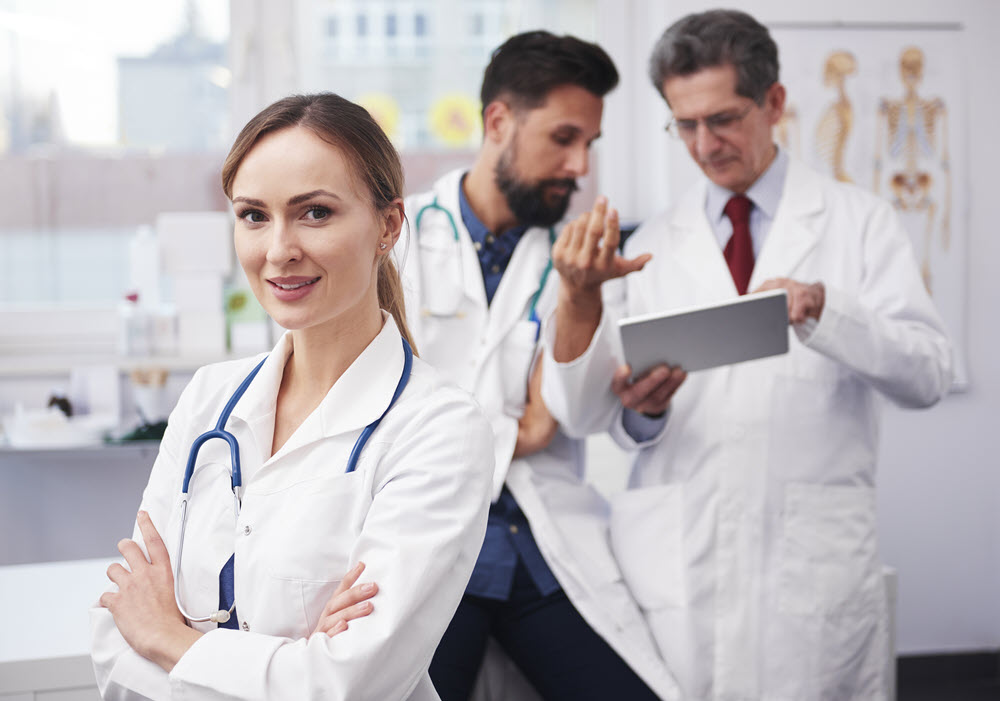A female doctor smiles into the camera as two male consulting in the background